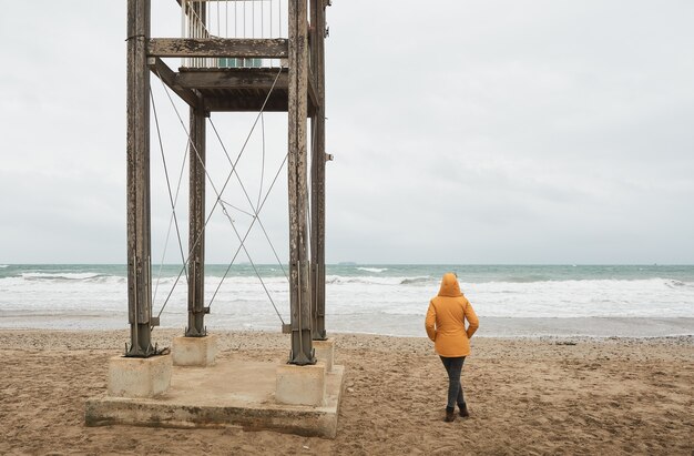 Fille en imperméable jaune regardant la mer. Marchez le long de la plage par mauvais temps. Personne marchant sur le sable à côté de la hutte de la garde côtière