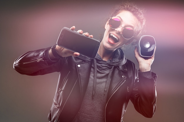 Photo fille impudente dans des verres avec un haut-parleur portable et un téléphone sur un mur noir avec effet lueur