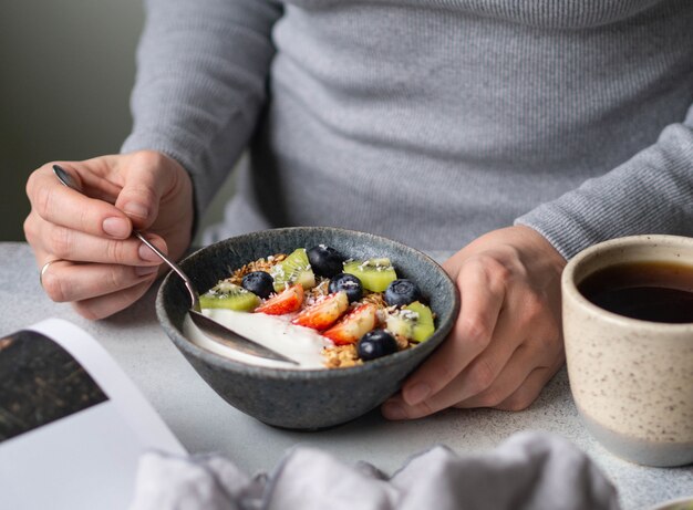 Fille incertaine en robe grise à la table avec petit déjeuner. Bol avec yaourt, granola et baies et tasse de café noir et magazine ouvert sur table grise