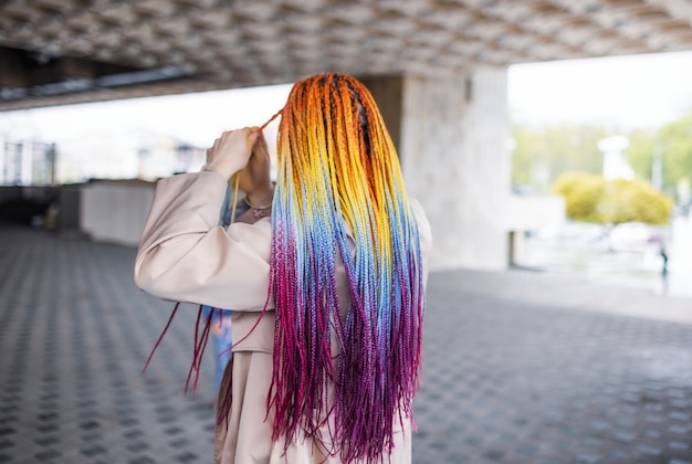 Fille inconnue à la mode dans une élégante veste beige avec des tresses africaines multicolores. Debout, dos à la caméra sur fond de rue de la ville, il redresse soigneusement ses tresses.