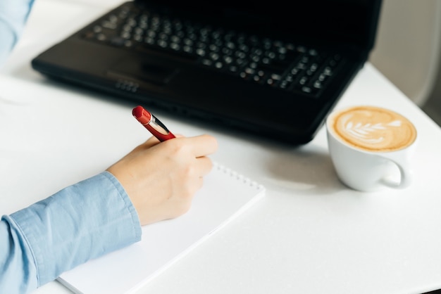 Une fille indépendante en chemise bleue écrit dans un cahier, sur le bureau se trouve un ordinateur portable noir et un café