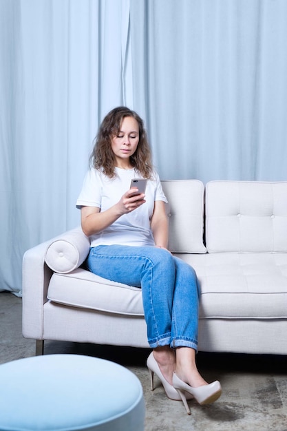 Une fille en jean bleu et un t-shirt blanc regarde le téléphone tout en étant assise sur un canapé de couleur claire