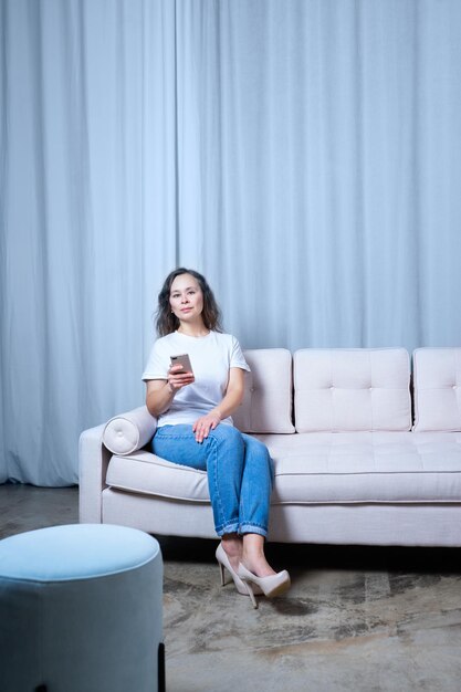 Une fille en jean bleu et un t-shirt blanc regarde le téléphone tout en étant assise sur un canapé de couleur claire