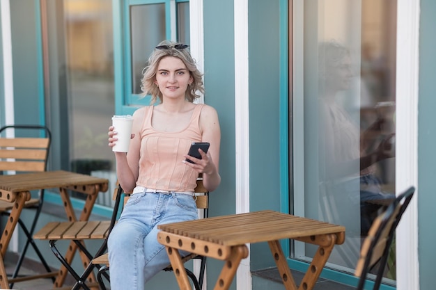 Une fille en jeans bleus est assise près d'un café avec un téléphone et une tasse de café en été