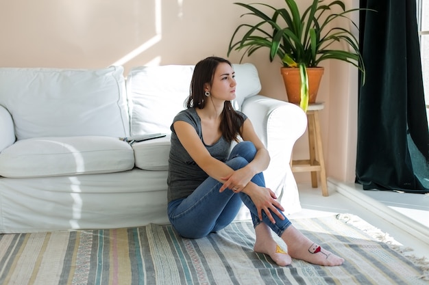 Fille en jeans est assise sur le sol, près du canapé, dans une pièce lumineuse.