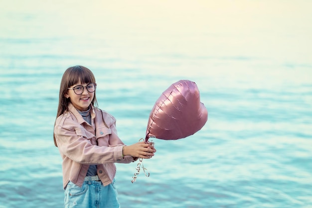 Une fille en jeans et une veste rose tenant un ballon au bord de la mer au coucher du soleil