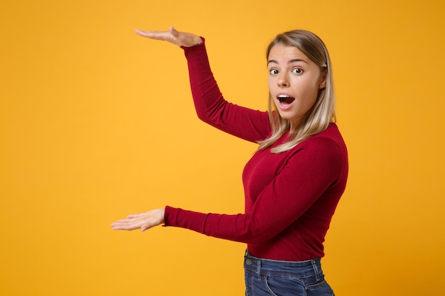 Fille jeune femme blonde choquée dans des vêtements décontractés posant isolé sur fond de mur orange jaune. Concept de style de vie des gens. Maquette de l'espace de copie. Gestes démontrant la taille avec un espace de travail vertical.