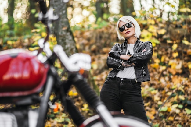 Fille jolie biker blonde à lunettes de soleil avec moto rouge sur la route dans la forêt