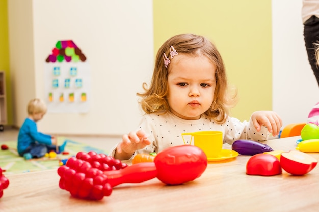 Une Fille Joue Avec Des Articles Ménagers Pour Enfants Et Des Fruits Artificiels à Table. équipement De Cuisine