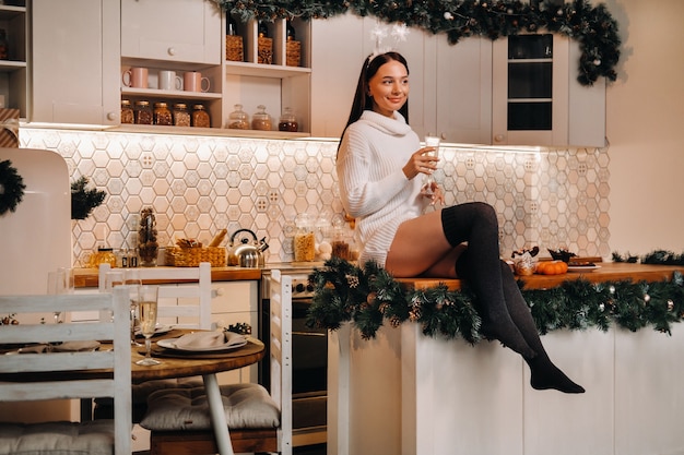 Une fille le jour de Noël est assise sur la table de la cuisine et tient une coupe de champagne. Femme le soir du nouvel an avec du champagne