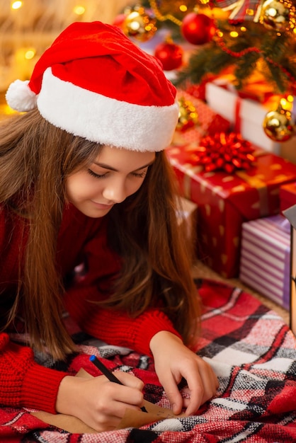 Fille joyeuse en bonnet de Noel assis près de l'arbre de Noël et des boîtes à cadeaux écrivant une lettre au Père Noël Concept de rêver une enfance heureuse et des vacances d'hiver