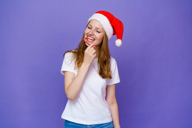 Une fille joyeuse en bonnet de Noel mange une sucette et rit en tshirt blanc jeune femme caucasienne e...