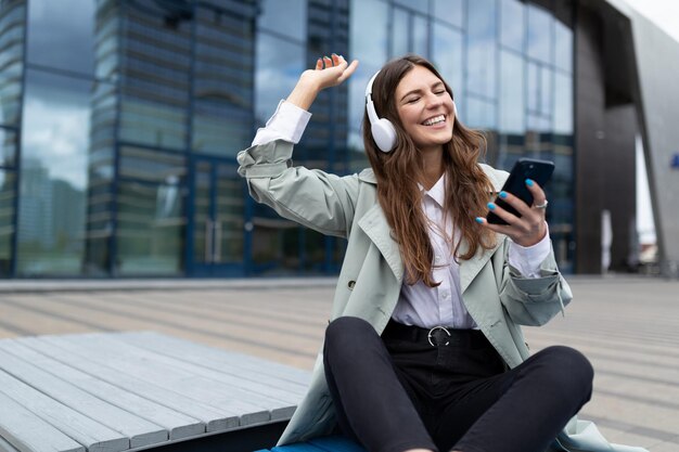 Une fille joyeuse dans les écouteurs est assise les jambes croisées sur un banc avec un téléphone dans les mains et danse