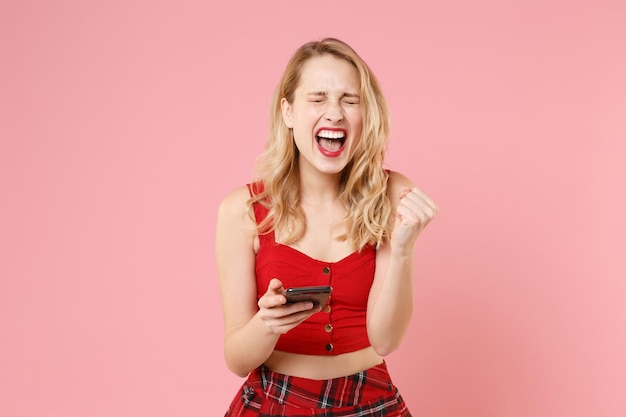 Fille joyeuse jeune femme blonde en vêtements sexy rouges posant isolé sur fond rose pastel. Concept de style de vie des gens. Maquette de l'espace de copie. À l'aide d'un téléphone portable en tapant un message sms, en faisant le geste du gagnant.