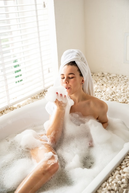 Une fille joyeuse joue avec de la mousse en prenant un bain au spa. Elle s'allonge dans la salle de bain et rit