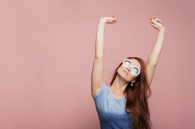 Une fille joyeuse en lunettes de soleil rondes bleues se réjouit et danse sur un mur rose. Heureuse jolie femme part en vacances en été chaud