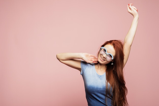 Une fille joyeuse en lunettes de soleil rondes bleues se réjouit et danse sur un mur rose. Heureuse jolie femme part en vacances en été chaud