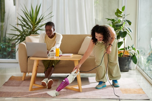 Une fille joyeuse passe l'aspirateur sur un tapis lorsque son colocataire étudie pour un examen