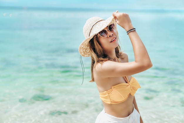 Fille joyeuse porte un bikini et un chapeau blanc posant émotionnellement avec les vagues de la mer et l'horizon en arrière-plan. Jolie femme à lunettes de soleil et tenue orange debout sur une plage de sable sur fond de mer bleue