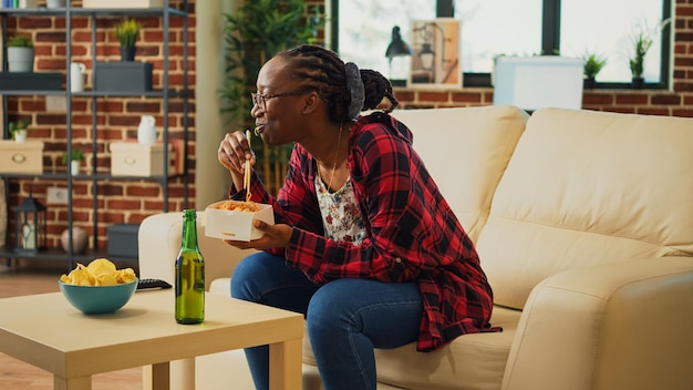 Fille joyeuse servant des nouilles dans une boîte de livraison, utilisant des baguettes pour manger de la nourriture asiatique et regarder un film d'action à la télévision. Femme appréciant le dîner du restaurant à emporter, s'amusant avec la télévision.