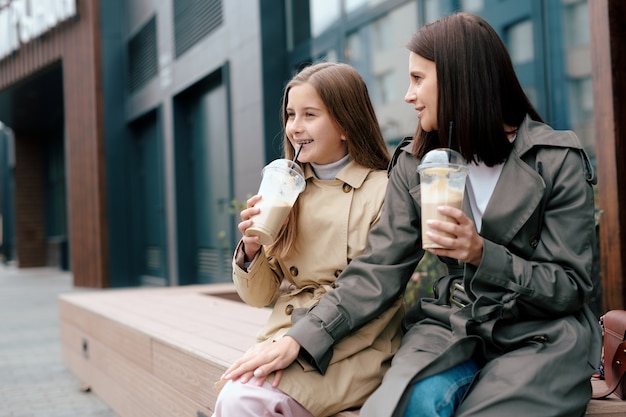 Fille joyeuse en trench en sirotant un cocktail tout en passant du bon temps avec sa mère et en discutant en milieu urbain