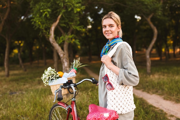 Fille joyeuse avec vélo et panier plein de fleurs sauvages et de fruits regardant joyeusement à huis clos passer du temps dans le parc