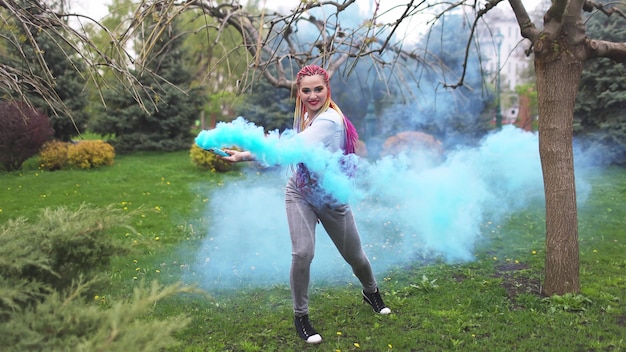 Une fille joyeuse vêtue d'une chemise et d'un jean avec des tresses arc-en-ciel lumineuses et un maquillage inhabituel. Elle danse cachée dans une épaisse fumée artificielle bleue