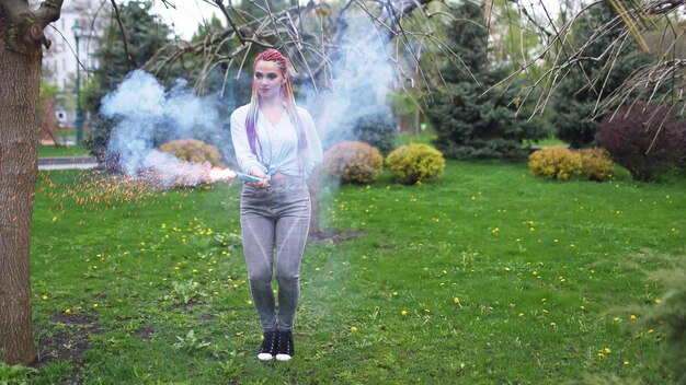 Une fille joyeuse vêtue d'une chemise et d'un jean avec des tresses arc-en-ciel lumineuses et un maquillage inhabituel. Elle danse en se cachant dans une épaisse fumée artificielle bleue sur fond de parc printanier