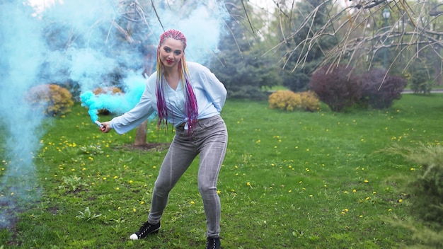 Une fille joyeuse vêtue d'une chemise et d'un jean avec des tresses arc-en-ciel lumineuses et un maquillage inhabituel. Elle danse en se cachant dans une épaisse fumée artificielle bleue sur fond de parc printanier