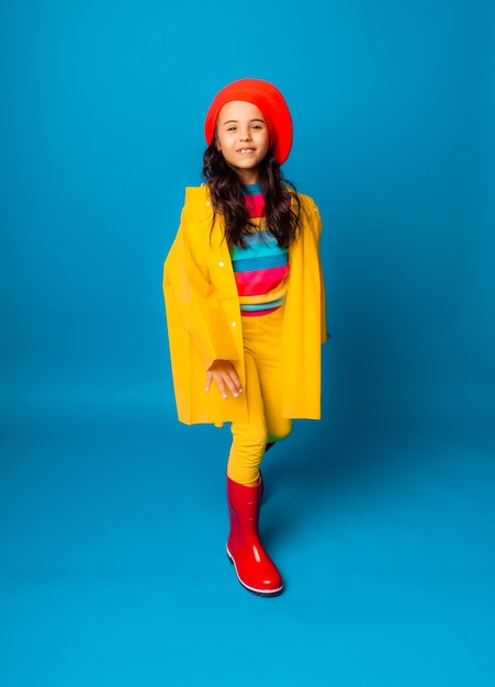 Une fille joyeuse vêtue d'un imperméable jaune, d'un béret rouge et de bottes en caoutchouc saute les bras tendus et regarde la caméra.