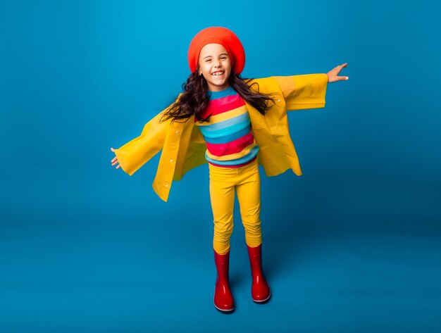 Une fille joyeuse vêtue d'un imperméable jaune, d'un béret rouge et de bottes en caoutchouc saute les bras tendus et regarde la caméra.