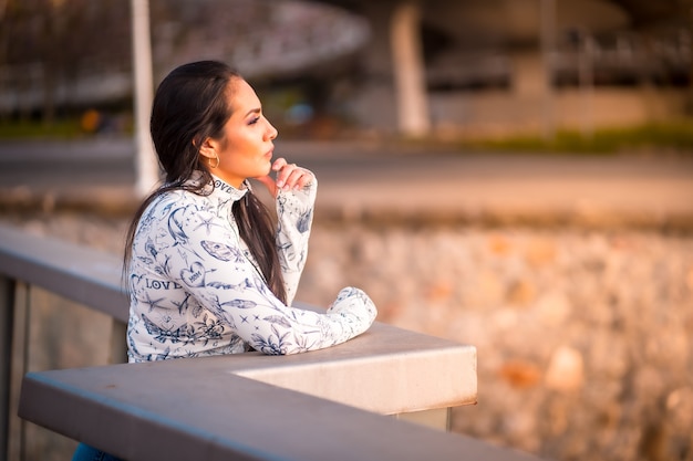 Une fille latine brune dans la ville au coucher du soleil en regardant la rivière sur un pont