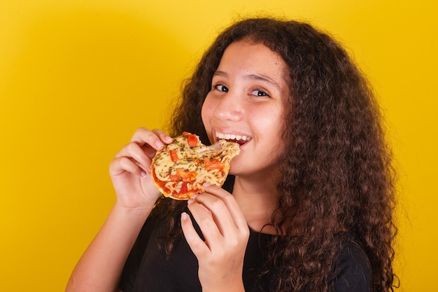 Fille latino-américaine brésilienne pour les cheveux afro fond jaune manger et mordre de délicieuses mini-pizza pizza margarita pizza fromage qui s'étend du fromage