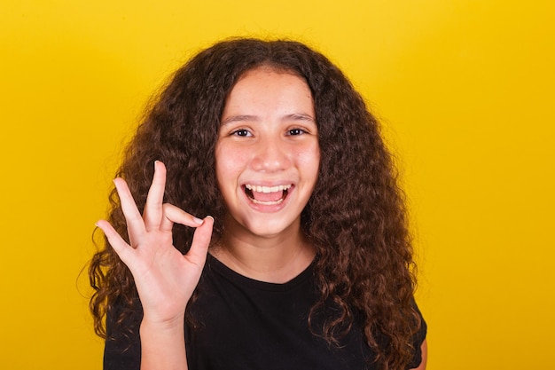 Fille latino-américaine brésilienne pour les cheveux afro fond jaune souriant faisant signe ok avec les doigts droit correct grand