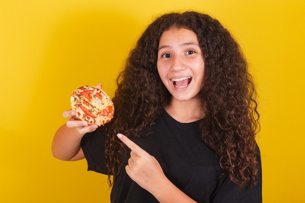 Fille latino-américaine brésilienne pour les cheveux afro fond jaune souriant suggérant une indication de suggestion pointant vers une mini pizza pizza pizza margarita fromage qui s'étend du fromage