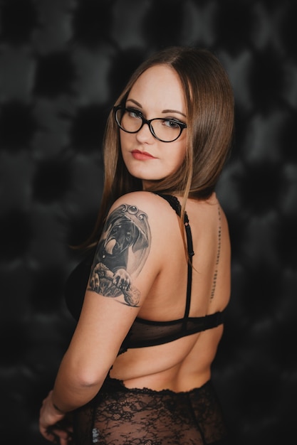 Fille en lingerie sexy noire et lunettes avec un tatouage sur son bras posant sur un mur noir