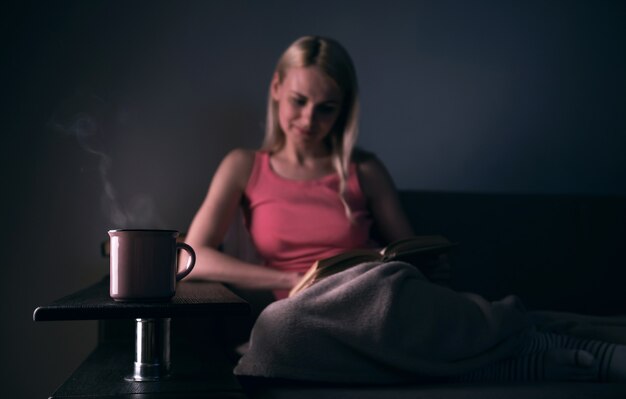 Fille lisant avec enthousiasme un livre maison à succès sous un tapis sur un canapé confortable avec une lampe