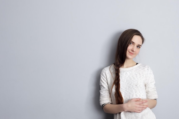 Fille avec une longue tresse debout devant le mur gris et regardant avec impatience modestement les mains jointes sur le ventre