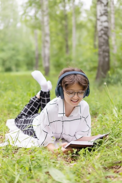 Une fille avec des lunettes et des écouteurs souriante lit un livre sur un pré vert pendant la journée d'été, vacances pour enfants