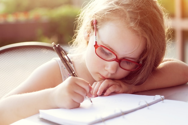 Fille avec des lunettes écrit dans un cahier. fond doux ensoleillé. faire ses devoirs à l'extérieur