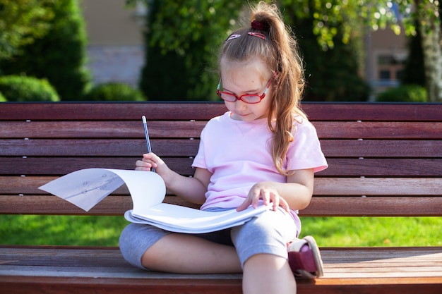 Fille avec des lunettes écrit et dessine dans un cahier à l'extérieur. devoirs dans la nature.