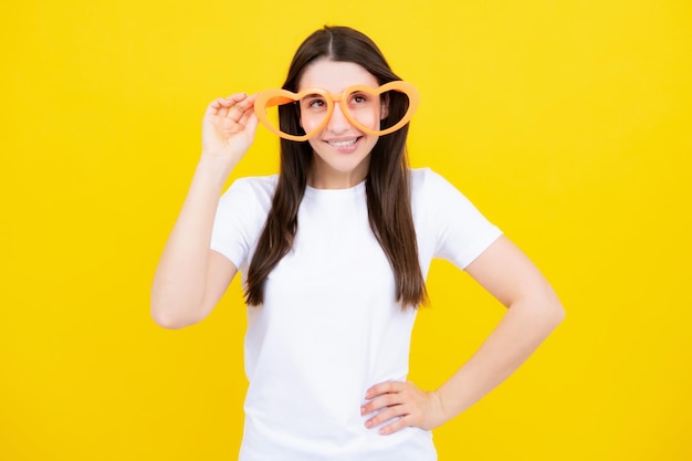 Fille avec des lunettes en forme de coeur drôle et souriant sur fond de studio jaune