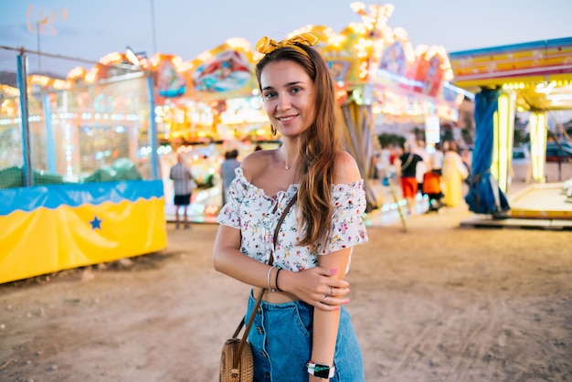 Fille à lunettes rondes cheveux bruns dans une jupe en jean et un sac à main rond sur le fond d'un parc d'attractions