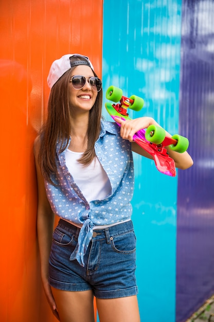 Fille à lunettes de soleil et casquette debout contre le mur coloré.