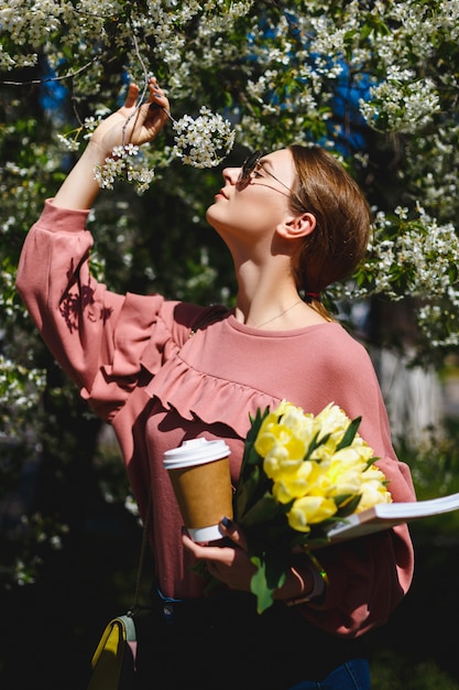 Fille à lunettes de soleil et un pull rose avec un bouquet de tulipes jaunes et de café parmi les arbres en fleurs