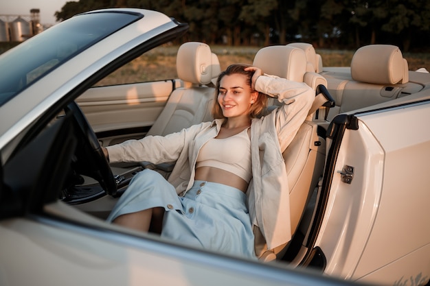 Une fille de luxe glamour aux cheveux blonds souriante assise sur une décapotable blanche. Jeune femme réussie s'asseyant dans sa voiture blanche