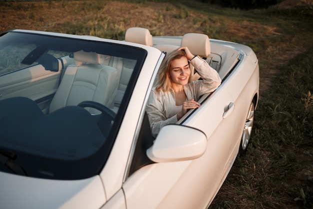 Une fille de luxe glamour aux cheveux blonds souriante assise sur une décapotable blanche. Jeune femme réussie s'asseyant dans sa voiture blanche