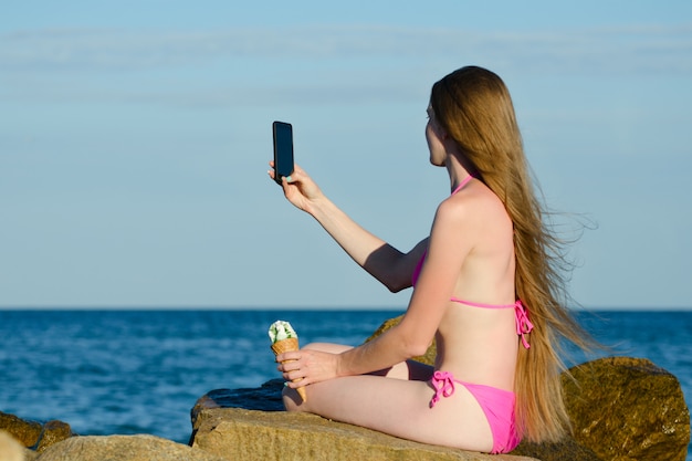 Fille en maillot de bain avec de la glace à la main sur la plage rocheuse fait selfie téléphone contre la mer