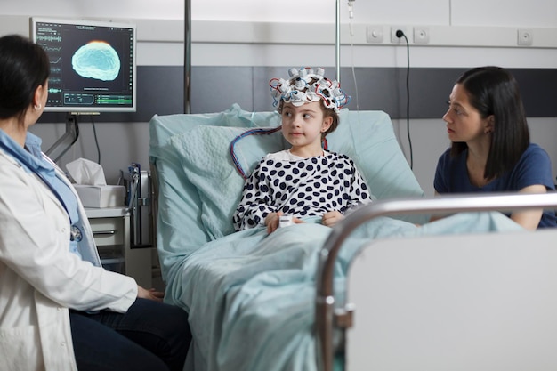 Une fille malade hospitalisée portant un casque EEG se reposant dans le lit du patient pendant que le médecin analyse l'état du cerveau. Spécialiste en neurologie examinant l'état de la maladie neurologique d'un enfant malade.