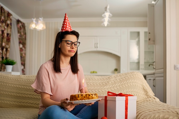 Une fille malheureuse portant un chapeau le jour de son anniversaire avec un gâteau aux chandelles est seule dans la pièce.